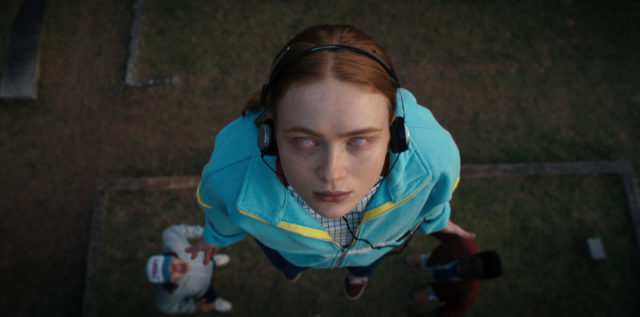Colored photo of Max Mayfield, played by Sadie Sink, levitating off the ground wearing earphones and a blue sweater.
