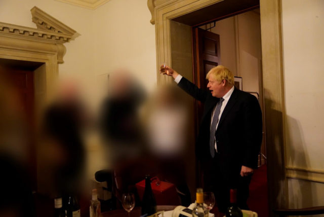 Colored photo of Boris Johnson raising a glass at a party beside three blurred figured.