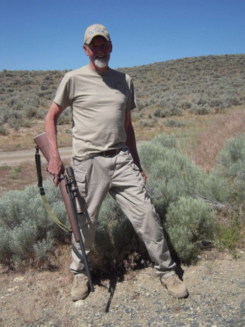 Chuck Mawhinney wearing all beige clothing wearing a hat, holding a rifle.
