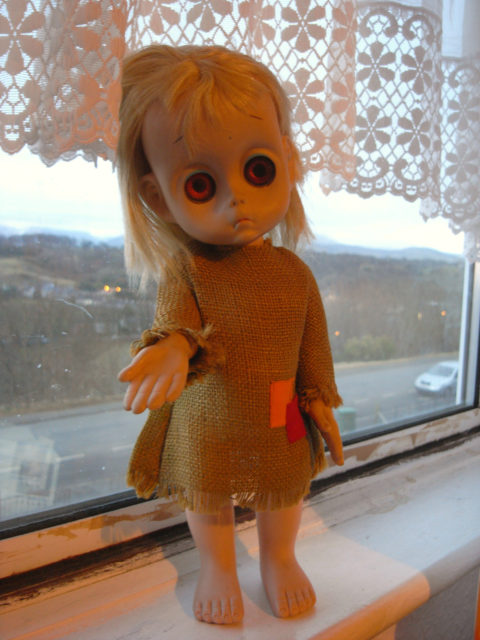 Colored photo of a Little Miss No Name doll standing on a window ledge.
