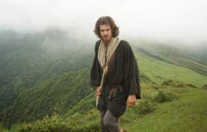 Andrew Garfield with long hair and a beard wearing a robe walking through a green mountain with fog in the background.