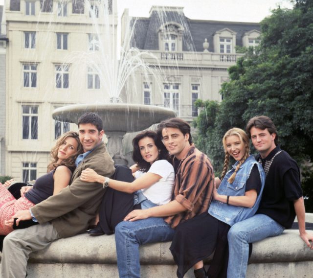 Colored photo of the cast of friends sitting on the edge of a fountain. 