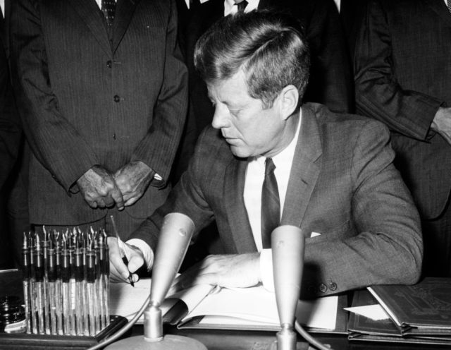 Black and white photo of John F. Kennedy signing the Partial Nuclear Test Ban Treaty while wearing a suit and looking down at the document.
