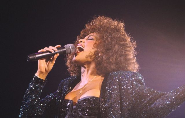 Whitney Houston in a sparkling black top, signing into a microphone. 
