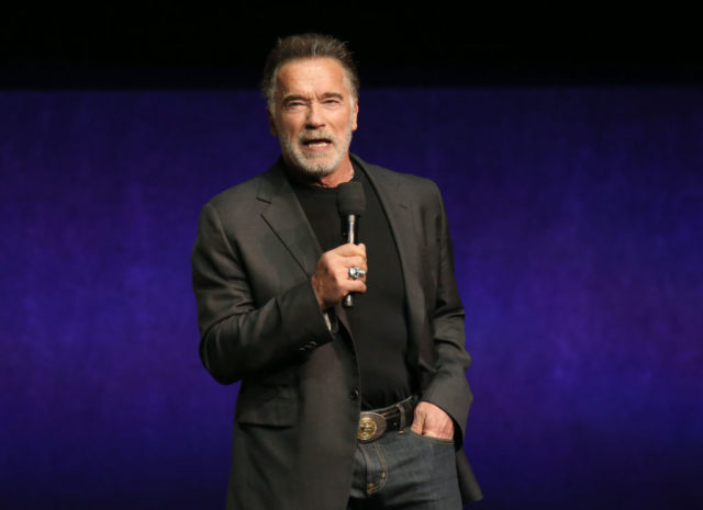 Colored photo of Arnold Schwarzenegger wearing a black shirt and jacket with jeans, standing with a microphone in front of a purple background.