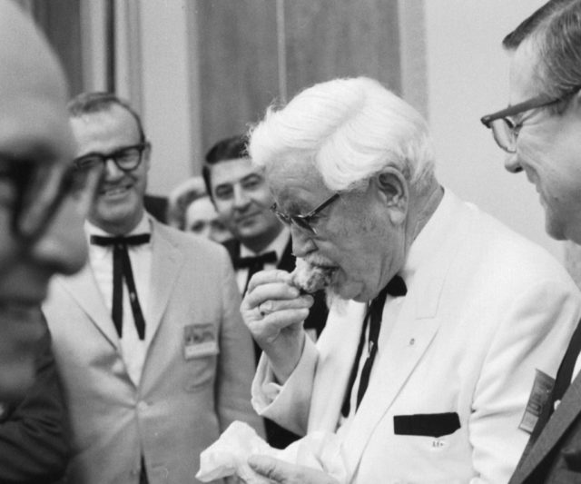 Group of people watching Colonel Sanders eating a piece of fried chicken