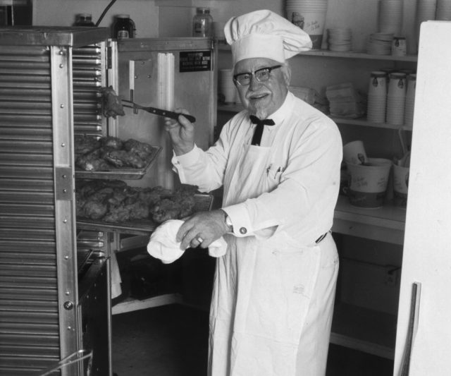 Colonel Sanders standing next to a rack of fried chicken