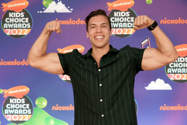 Joseph Baena flexing his muscles in a black collared t-shirt while smiling at the camera.