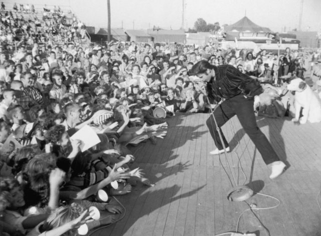 Black and white photo of Elvis signing and dancing while holding a microphone on a stage in front of a cheering crowd.