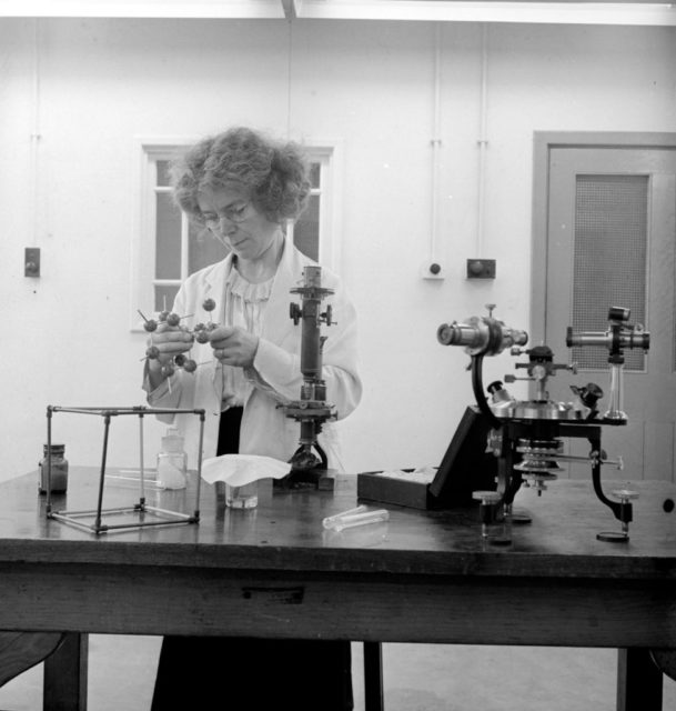 Black and white photo of Kathleen Lonsdale standing behind a lab bench looking down at a molecule model.