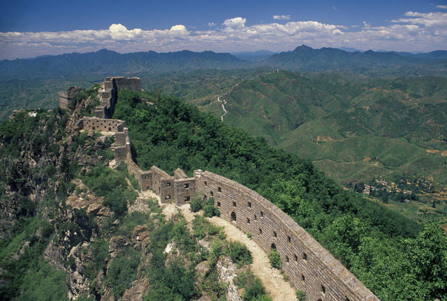 Thin segment of the Great Wall of China in a green mountainous area stretching off in the distance.