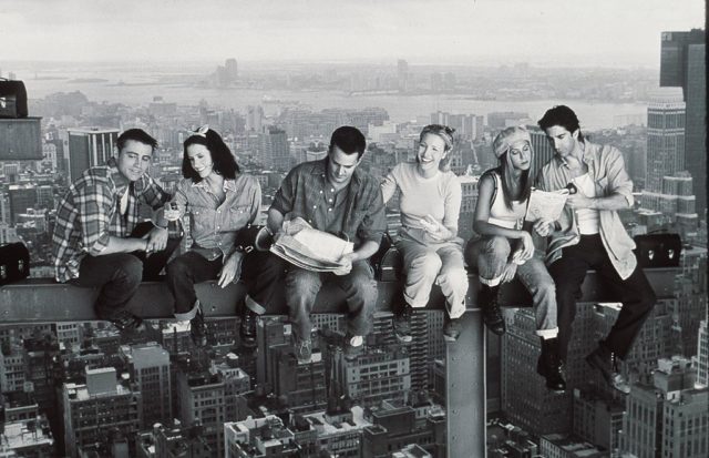 Black and white photo of the cast of friends sitting on a scaffolding beam above the city. 