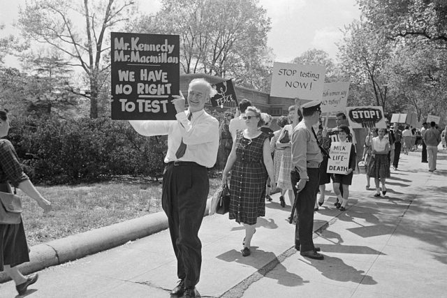 Black and white photo of a group of protestor walking on a sidewalk holding signs against nuclear testing.