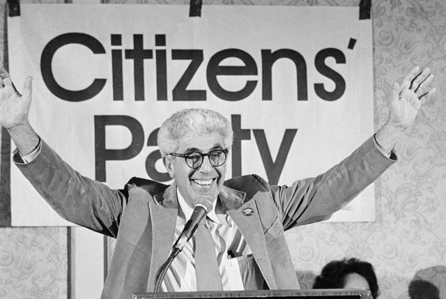 Black and white photo of Dr. Barry Commoner in front of a podium with his arms in the air smiling at the camera.