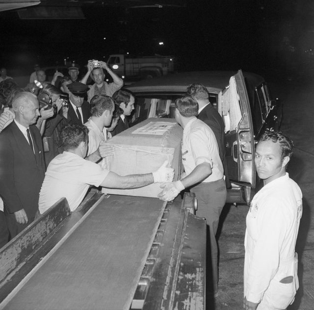 Funeral home workers loading a wooden casket onto an airplane conveyor belt