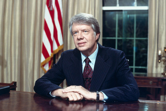 Colored photo of Jimmy Carter in a suit sitting at his desk in the White House.