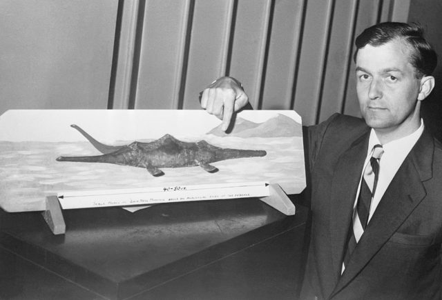 Black and white photo of a man in a suit pointing at a hand drawn depiction of the Loch Ness Monster.