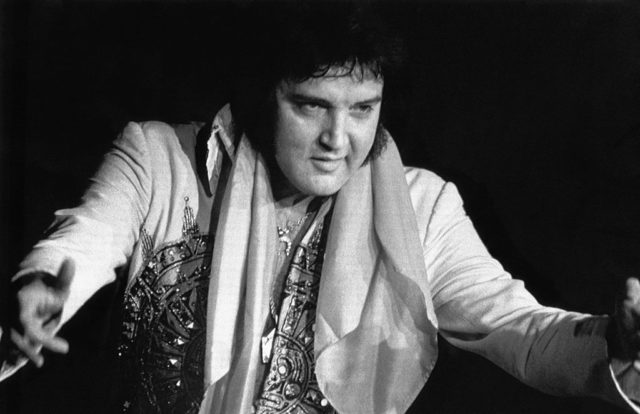 Black and white photo of Elvis Presley with his arms open wearing a costume on stage.