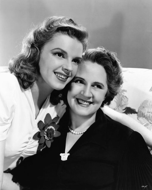 Judy Garland in a white shirt with a flower pinned on it leaning over to hug her mother, Ethel Gumm.