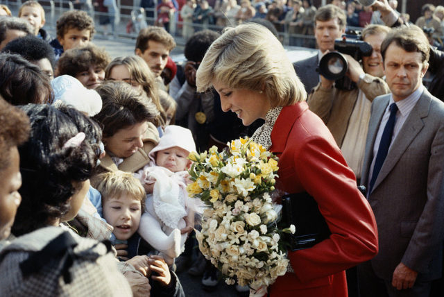 Colored photo of Princess Diana in a red coat accepting flowers from a crowd. 