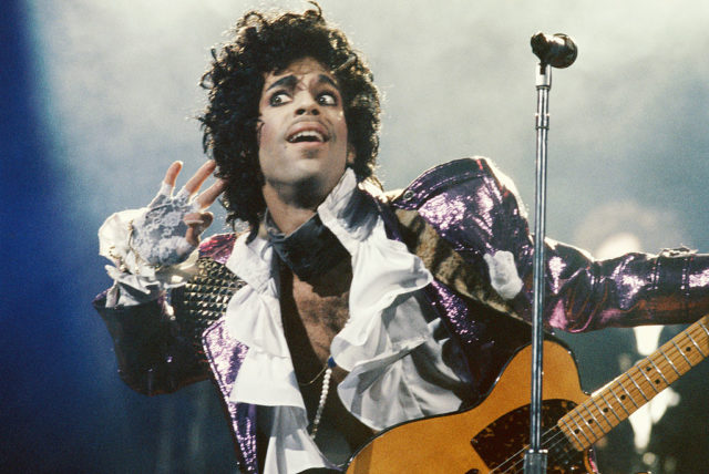 Prince holding a guitar in front of a microphone stand holding his hand up to his ear wearing a ruffled shirt.