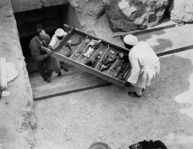 Black and white photo of two men carrying a tray of objects up stairs leading out of an underground tomb.