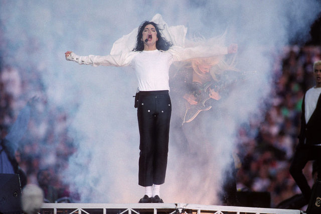 Michael Jackson in a white shirt and black pants throwing his arms out to the side while singing on stage.