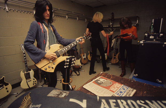 Joe Perry, Tom Hamilton and Joey Kramer standing among a number of guitars