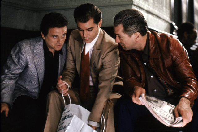 Colored movie still of Ray Liotta, oe Pesci, and Robert De Niro standing around a paper bag.