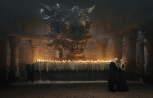 Colored movie still of Milly Alcock and Paddy Considine standing in front of a table with melting candles, with a dragon head above it.