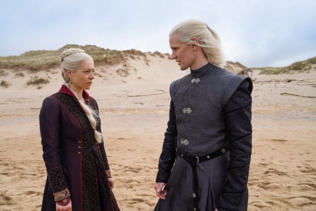 Colored movie still of Matt Smith and Emma D'Arcy standing on a beach wearing Mediaeval style clothing. 