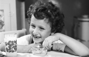 Black and white photo of a young girl sitting at a table with a glass of milk smiling with her front teeth missing.