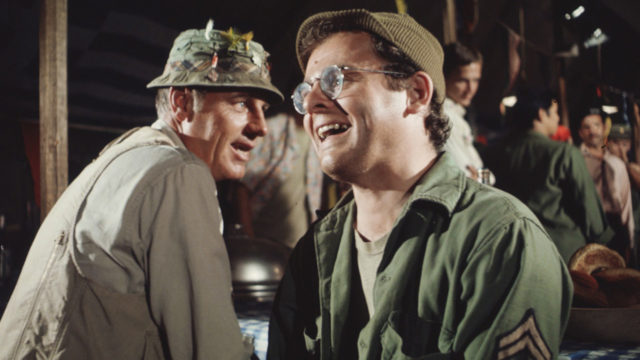 Colored movie still of McLean Stevenson in a tan vest and fishing hat, and Gary Burghoff in a green hat and green army jacket.