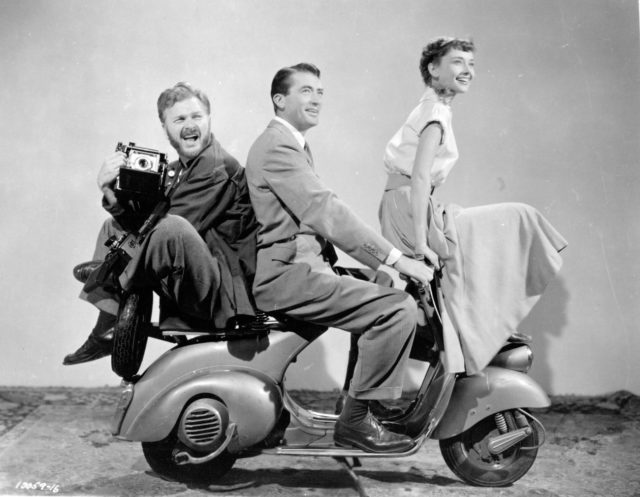 Black and white photo of Gregory Peck, Audrey Hepburn, and Eddie Albert riding a moped. 