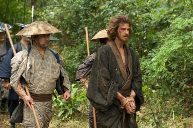 Andrew Garfield in character wearing a robe with long hair and a heard being escorted by a group of Japanese men in the movie 'Silence.'