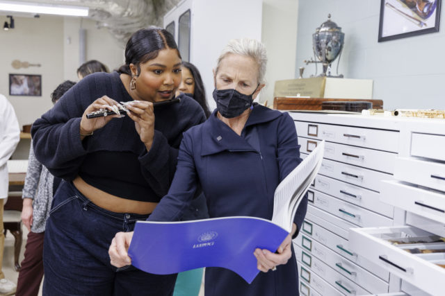 Lizzo holding a flute and reading sheet music held by another woman