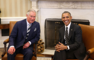 King Charles III in a blue suit sits beside President Barrack Obama wearing a black suit.