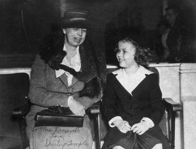 Eleanor Roosevelt wearing a hat and holding a handbag sits beside Shirley Temple who smiles up at her.
