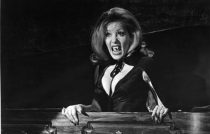 Female vampire standing up in a coffin bearing her teeth at the camera.