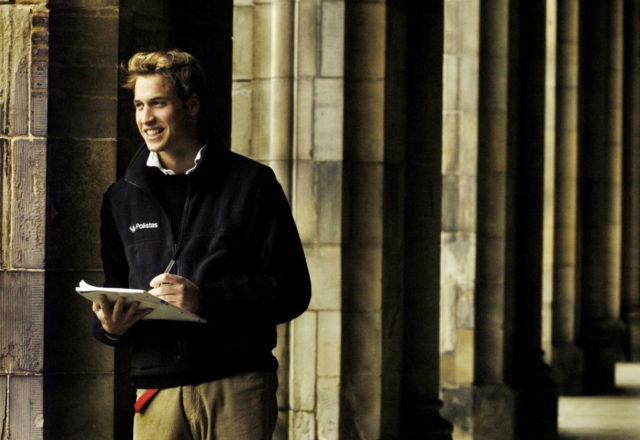 Prince William leaning against a concrete pillar holding a pen and paper.