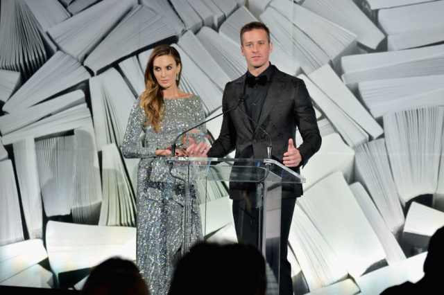 Armie Hammer in an all black suit standing behind a glass podium beside Elizabeth Chambers in a floor length sparkling gown.