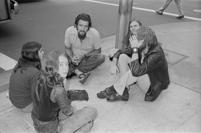 Group of Manson Family members sitting outside on the sidewalk. 