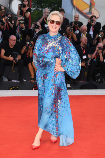 Meryl Streep in a blue dress, red heels, and sunglasses on a red carpet. 