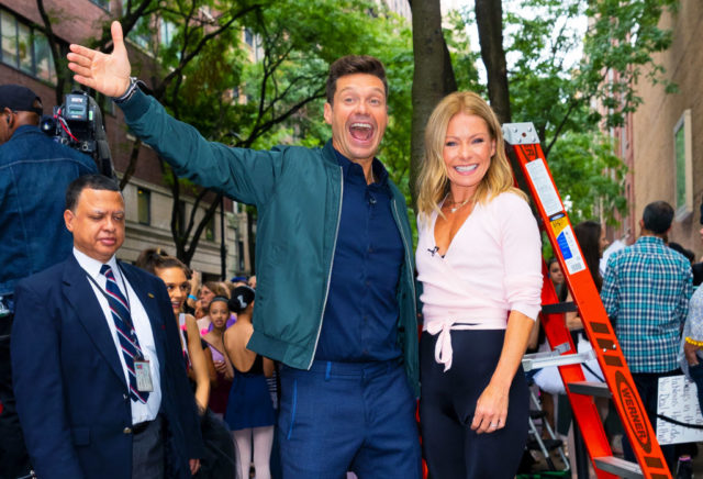 Ryan Seacrest in a jacket holding his arm out in enthusiasm, and Kelly Ripa smiling beside him.
