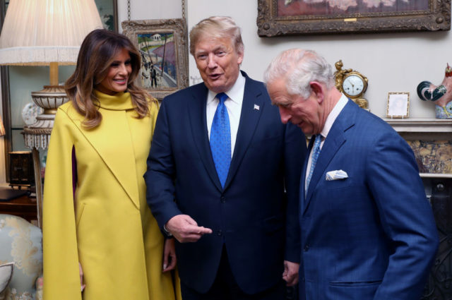 Melania and Donald Trump stand beside King Charles III, who tilts his head down while Trump points at him.