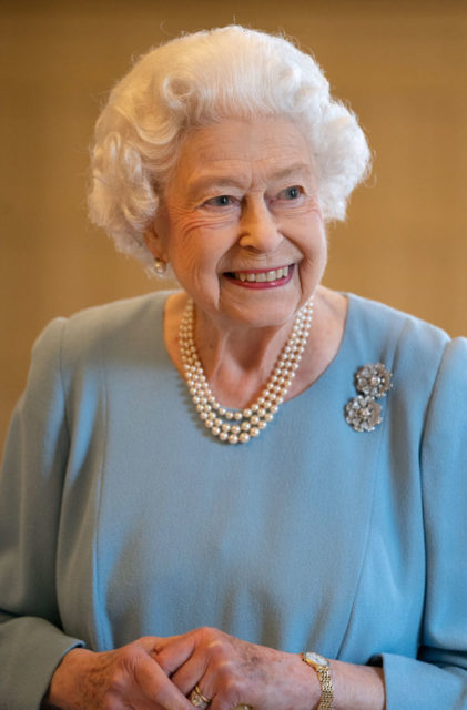 Queen Elizabeth II in a blue dress and pearls smiling at the camera.