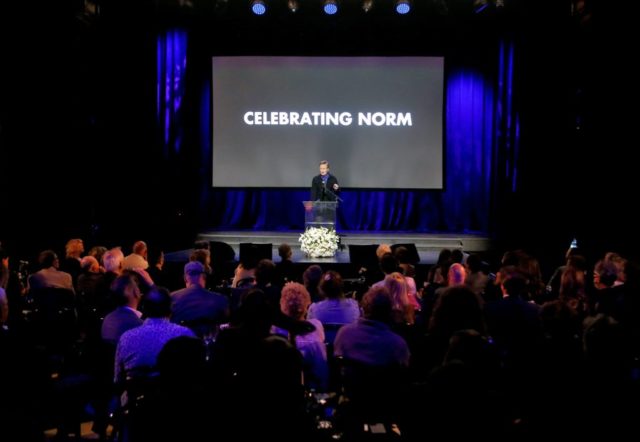 Group of people in the audience face a stage where Conan O'Brien speaks in front of a screen which reads "Celebrating Norm."