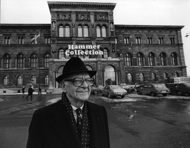 Black and white photo of Armand Hammer in a black coat and hat standing in front of a sign on a building reading "Hammer Collection."