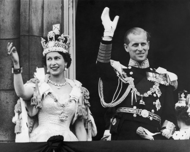 Black and white photo of Queen Elizabeth II in her coronation gown and crown wave on a balcony beside the Duke of Edinburgh. 
