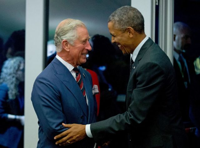 King Charles III in a blue suit and President Barrack Obama in a black suit shake hands.